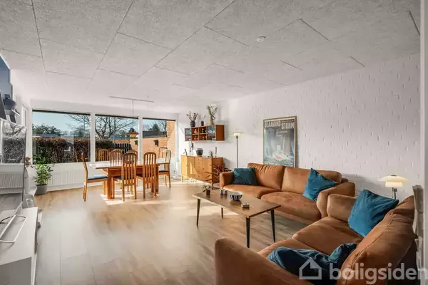 Brun sofa med blå puder står i en stue med lyst trægulv. Ved siden af ses et træspisebord med seks stole. Store vinduer giver udsigt til en have.