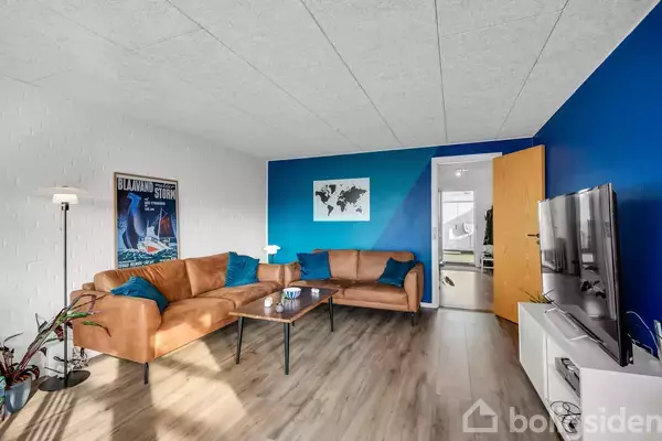 Brunt lædersofaarrangement med blå puder står i et moderne rum med blå accentvæg og trægulv. Et rundt sofabord er foran sofaen. Billedet på væggen viser et verdenskort. Vegetation til venstre.