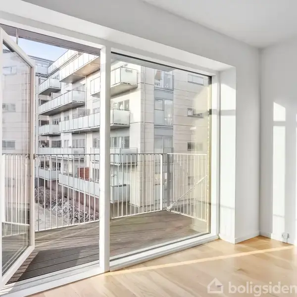 En stue har store glasdøre, som står åbne mod en altan med udsigt til moderne lejlighedsbygninger. Sollys strømmer ind og skaber skygger på trægulvet.