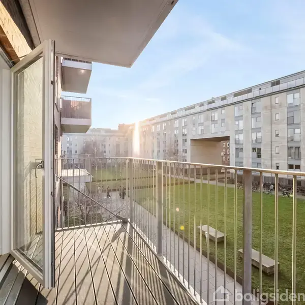 En åben dør fører til en altan med metalrækværk, hvor solen skinner over en grøn gårdhave omgivet af lejlighedsbygninger i baggrunden.