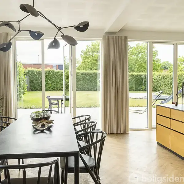 Sort spisebord med stole i spiseområde, omgivet af store vinduer og døre, der åbner ud til en grøn have. Moderne loftslampe og køkkenø med planter i baggrunden.