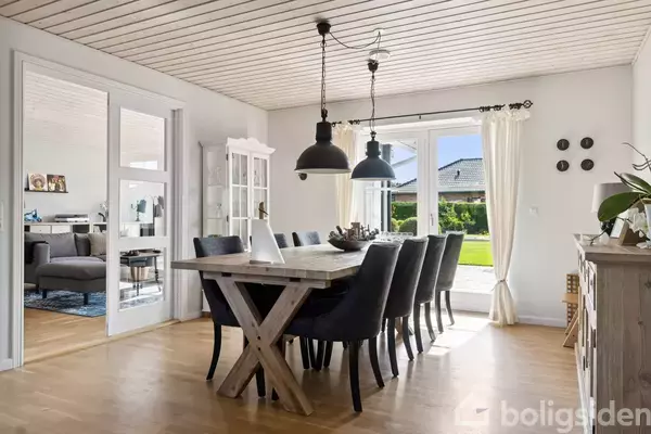 Spisebord med sorte stole står i lyst spisestue, oplyst af store vinduer med udsigt til have. Spisestuen er forbundet med en stue via glasdøre, med træloft og lyse gardiner.