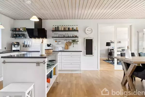 Køkken med hvidt interiør dominerer, mens hylder med krydderier og et stort køkkenbord skaber en arbejdszone. I baggrunden er der adgang til en stue med afslappet atmosfære.