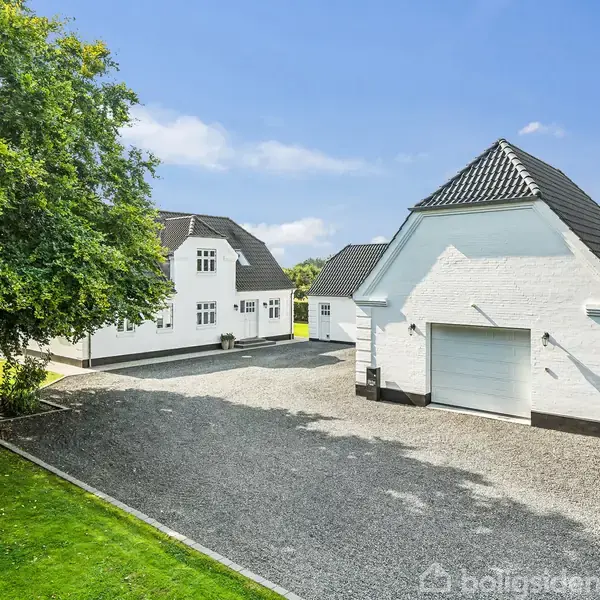 Hvidt hus og garage står adskilt; begge har mørke tegltage. Solen skinner, omgivet af grusbelægning og grøn græsplæne med træer. Klar blå himmel i baggrunden.