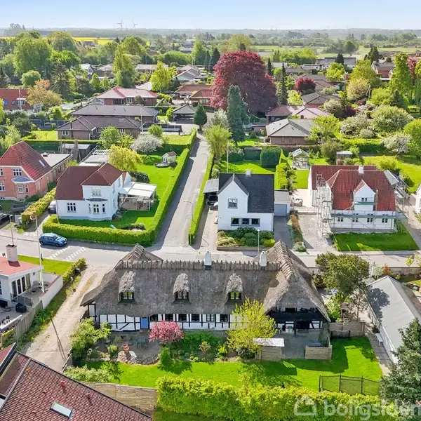 Stråtækt bindingsværkshus omgivet af frodige haver i et villakvarter. Flere moderne huse ses rundt omkring, og områderne er tæt beplantet med træer og buske.