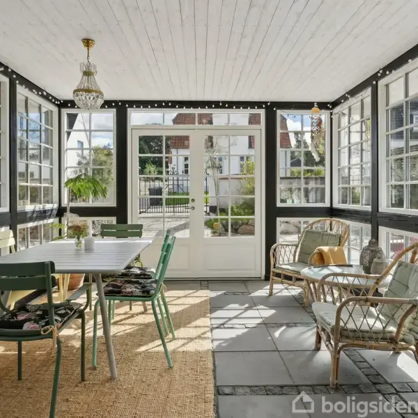 Spisestuemøbler står på en tæppebelagt veranda med glasvægge og hvide lofter. Sollys strømmer ind, og der er planter og udendørs udsigt.