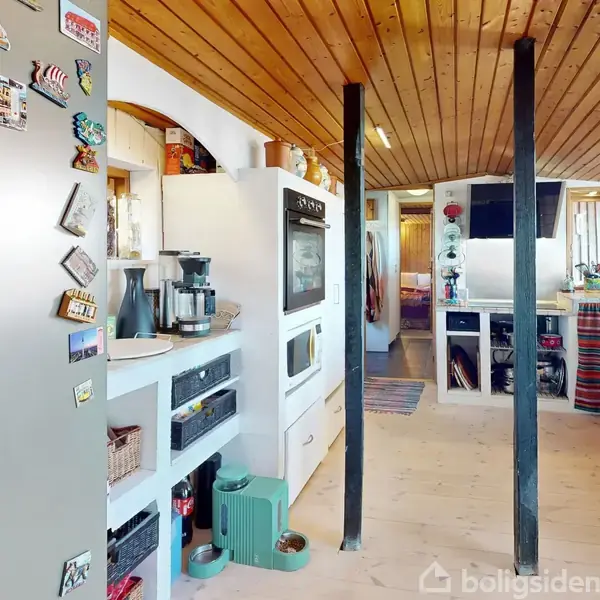 Et moderne køkken med en ovn indbygget i hvid skabsvæg. Køkkenmaskiner er placeret på en hylde. Rummet er lyst med træloft og farverige gardiner. Magneter pynter køleskabet.