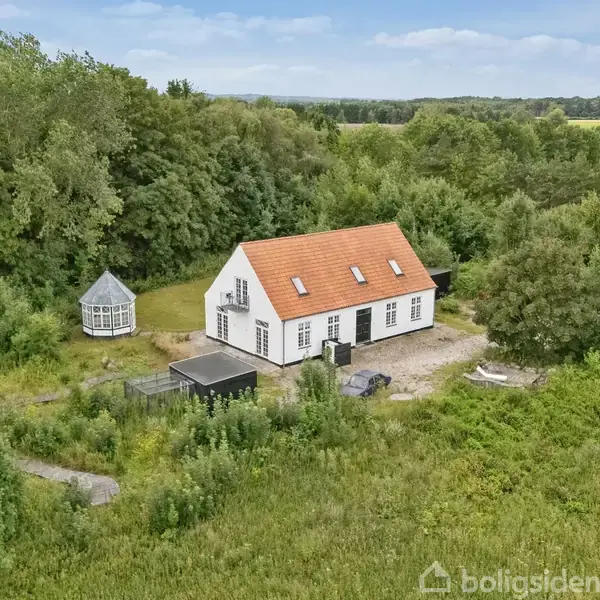 Et hvidt hus med rødt tag står omgivet af tæt skov og grønne marker. Der er et lille lysthus til venstre og en flisebelagt indkørsel foran huset.