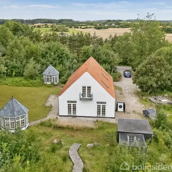 Fritliggende hvidt hus med rødt tag, omgivet af grøn skov og marker. To havepavilloner i nærheden. En grusvej fører til en parkeret bil ved siden af huset.