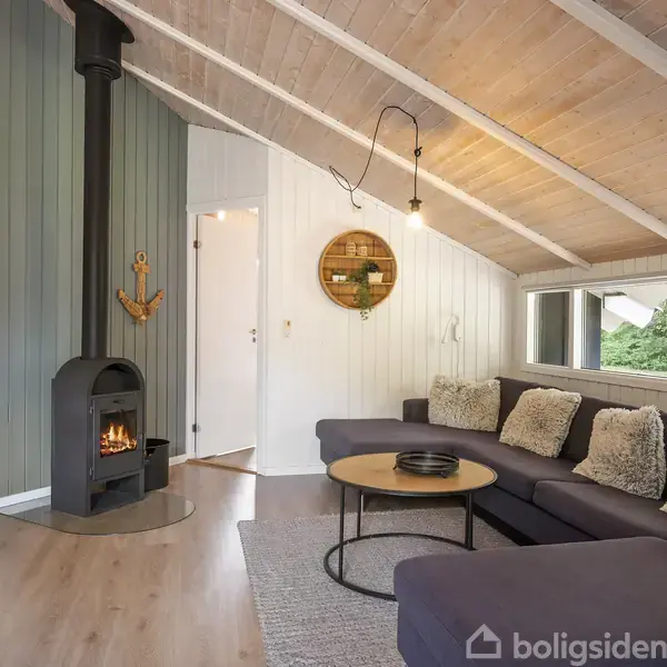 Brændeovn opvarmer stue med træbeklædte vægge, skrå loft. En stor sofa med puder står foran et rundt sofabord. Væggene har en dekorativ kroganker og en hylde.