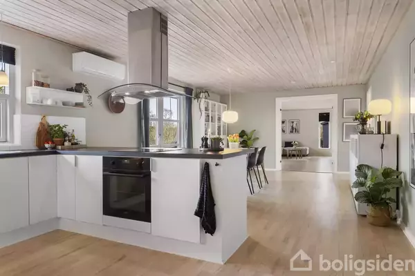 Et moderne køkken med hvidt skab og indbygget ovn. Der hænger et emfang over kogepladen. Rummet er lyst med træloft og gulv, og spisestue ses i baggrunden.