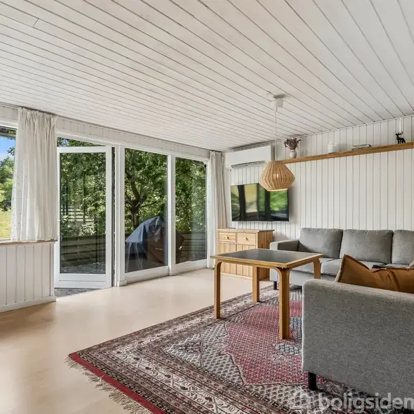 Grå sofa står i stuehjørnet på et mønstrede tæppe. Store vinduer viser udsigt til græsklædte bakker. En hylde med bøger og en lampe hænger over sofaen.