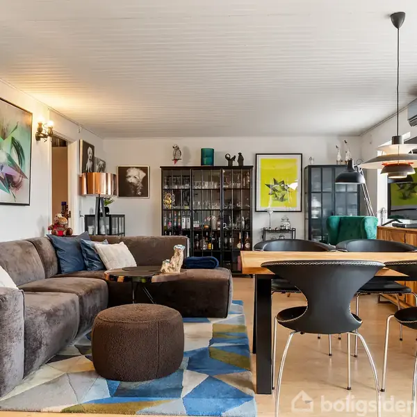 Brun sofa står i en stue med farverige puder. Spisebord til højre omgivet af moderne stole. Vægkunst og reol fylder baggrunden. Lysindfald fra vindue skaber en hyggelig atmosfære.