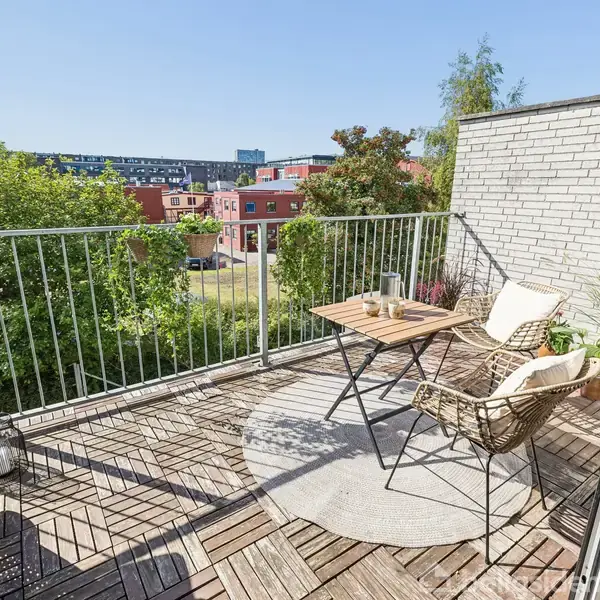 En lille altan med trælaminatgulv og et bord med to stole står i solen. En grill er placeret til venstre, og omgivelserne inkluderer beplantning og farverige bygninger i baggrunden.