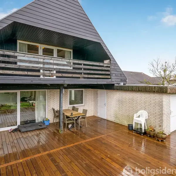 Hus med en stor overdækket terrasse. Træ-terrassen har møbler og planter, og ovenpå er der en altan med gelænder. Området er åbent og grønt med flere bygninger i nærheden.
