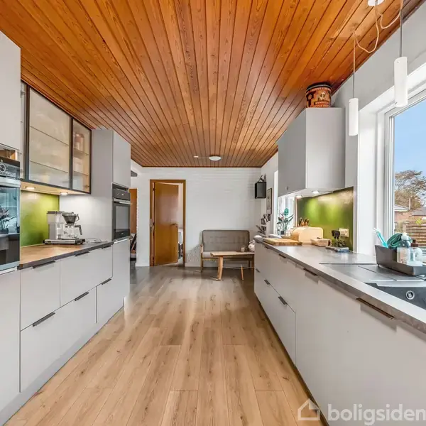 Køkken med moderne grå skabe og indbyggede ovne, i aktion med stilfuldt interiør. Træloft og trægulv skaber en varm atmosfære. Stor vindue giver udsigt til en gade.