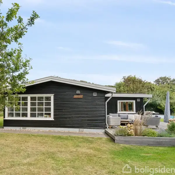Sortmalet, étplans hus med store vinduer står stille på en velplejet græsplæne. Til højre er en terrasse med møbler, omgivet af frodig vegetation og med træer i baggrunden.