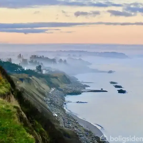 Klippe kystlinje omkranses af græs, mens tåge svæver over havet. Kysten strækker sig mod horisonten under en blå og lyserød himmel.