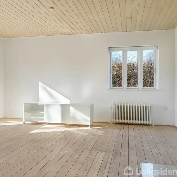 Rum med trægulv og hvide vægge. En brændeovn er tændt i hjørnet. Et skrivebord står foran et vindue med udsigt til buske udenfor.