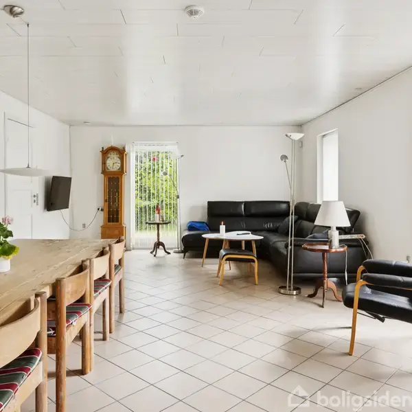 En stue med et langt træbord og stole til venstre. En sort sofa og lænestole står til højre. Et gulvur i baggrunden ved en dør med udsigt til grønne planter.