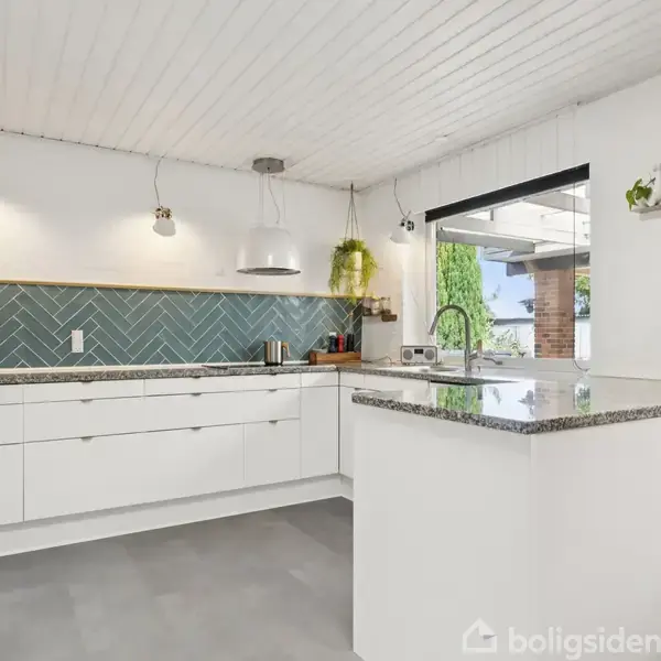 Køkken med hvide skabe og grå bordplader. Mikroovn og bøger står til venstre. Grønne fliser på væggen. En hylde med ur og blomster. Udsigt til terrasse gennem vindue.