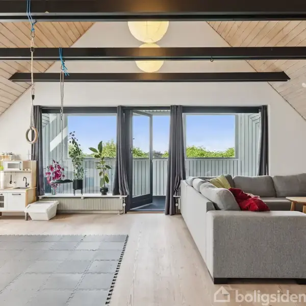 En lys stue med en grå hjørnesofa og legetæppe. Der hænger gymnastikringe fra loftet. Store vinduer åbner ud til en balkon med udsigt til grønne omgivelser.