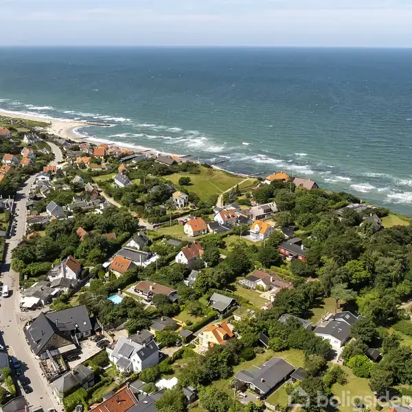 Kystby med små huse spreder sig langs en vej; bølger ruller ind mod den sandede kystlinje. Hussene er omgivet af frodig vegetation, og havet strækker sig ud i horisonten.