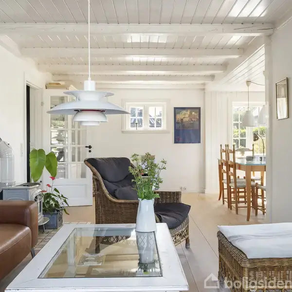 En stue med en lædersofa og glasbord, omgivet af flettede stole og spisebord. Lyse vægge med vinduer lader naturligt lys ind. Grønne planter tilføjer farve.