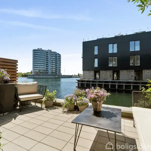 En terrasse med borde og stole har udsigt over en kanal med moderne bygninger på begge sider. Terrassen er omgivet af planter og blomster, der skaber en hyggelig atmosfære.