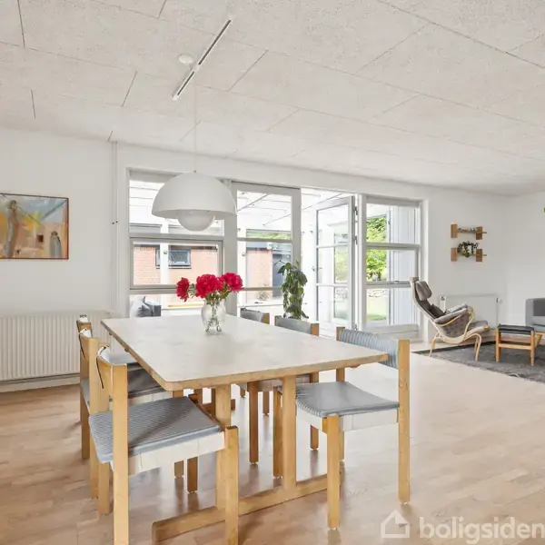 Spisebord med stole står i en lys stue. En vase med blomster pryder bordet. Baggrunden viser en sofa, en lænestol og store vinduer med udsigt til en terrasse.