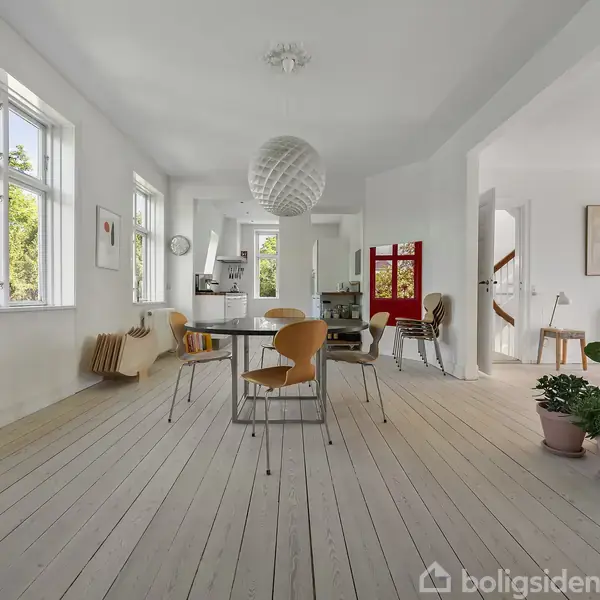 Spisebord med stole står i et rummeligt, lyst køkken-alrum. Et rundt loftlys hænger over bordet. Rummet har hvide vægge, trægulve, planter og kunstværker på væggene.