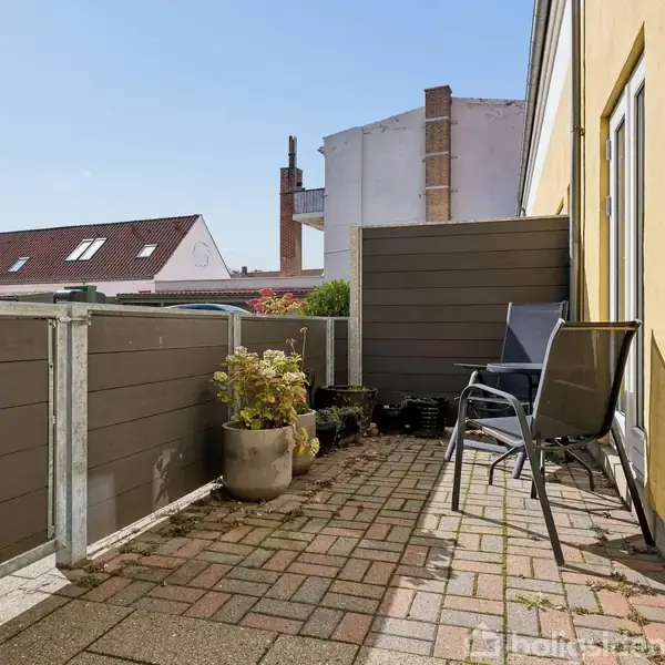 En lille, solbelyst terrasse har to stole og en pottet plante stående mod en gul væg. Omkranset af et lavt stakit med udsigt til hustage i baggrunden.