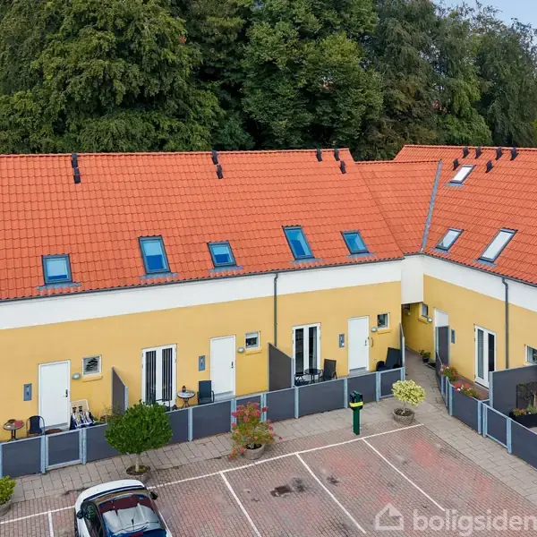 Rækkehuse med gule facader og røde tegltage står på række. Vinduer og døre åbner ud mod en gårdsplads med parkeringspladser. Skovområde ses i baggrunden.