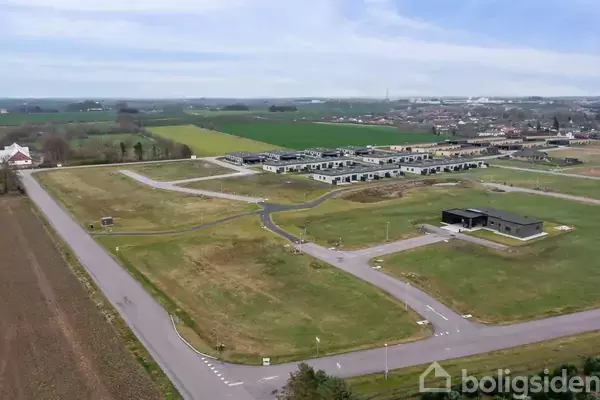 Luftfoto af et nybyggerkvarter med veje og ubebyggede grunde i en åben, grøn landskab. Et boligområde ses i baggrunden.