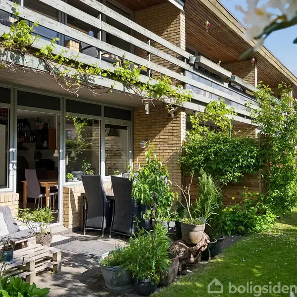En terrasse med træmøbler og planter står foran en murstensbygning med store vinduer. Terrassen er omgivet af frodig beplantning, og en græsplæne strækker sig langs bygningen.