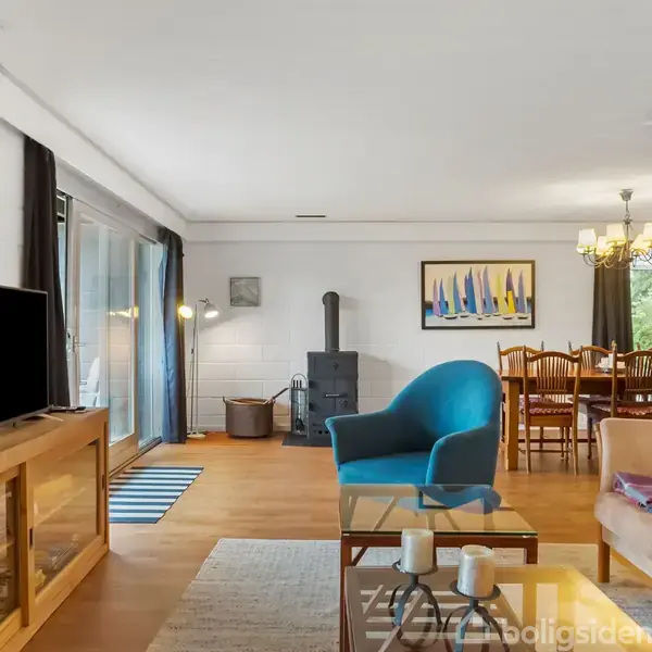 Stue med sofa, blå stol og tv-bord til venstre. Spisebord med stole og lysekrone i baggrunden. Parketgulv og vægkunst bidrager til en hyggelig atmosfære.