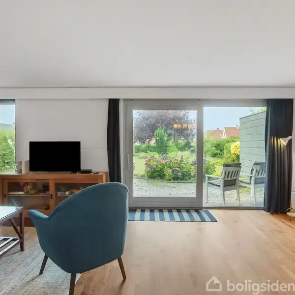 Stue med blå stol og sofabord, udsigt gennem glasdør til velholdt have med blomster og havemøbler. Vægge med lys indretning og træmøbler.