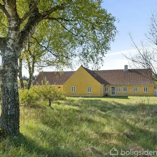 En lang, gul bygning med rødt tegltag står i en græsbevokset have, omgivet af træer og en skov i baggrunden på en solrig dag.