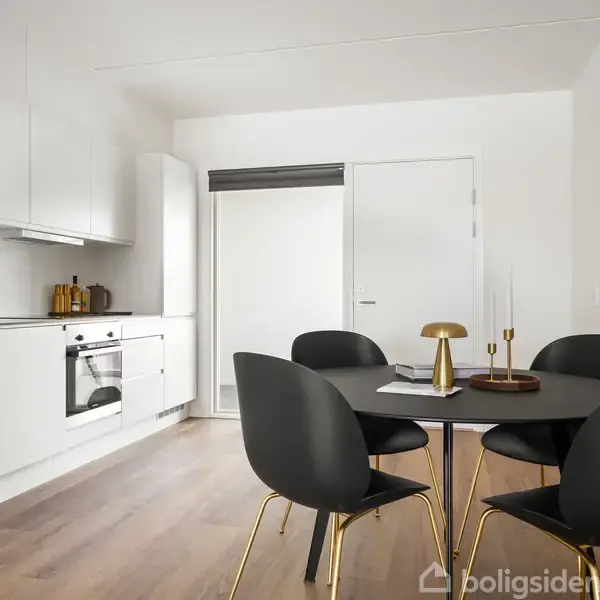 Et rundt spisebord med sorte stole står i et moderne køkken. Køkkenet har hvide skabe, en indbygget ovn og køleskab. Væggen har et maleri, og gulvet er træbelagt.