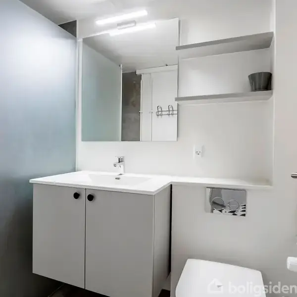 Et moderne badeværelse med hvidt skabsarrangement under vasken, en toilet til højre og hylder med dekorative elementer. Et stort spejl og bruseområde med matteret glas er til venstre.