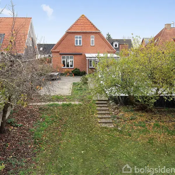 Et rødt murstenshus med en trekantet tagform står i en have. Træer og buske omgiver en græsplæne. Fliser danner et terrasseområde bag huset. Ingen tekst findes i billedet.