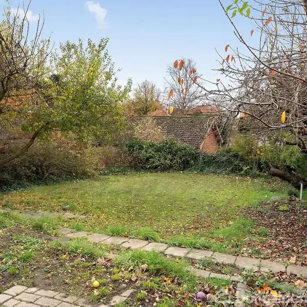 Græsplæne omgivet af buske og træer, nogle med efterårsblade, står stille i en have. Der er en murstensmur i baggrunden og nogle flisebelagte stier.