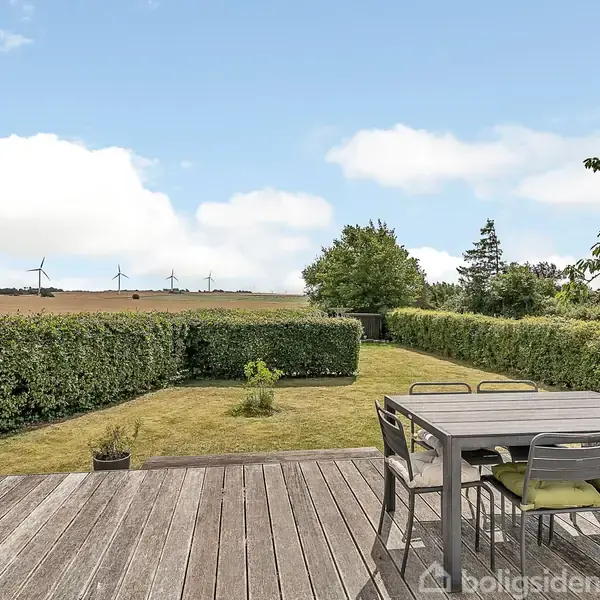 Terrassebord med stole; står stille; omgivet af grøn have og marker med vindmøller i baggrunden, under en blå himmel med hvide skyer.