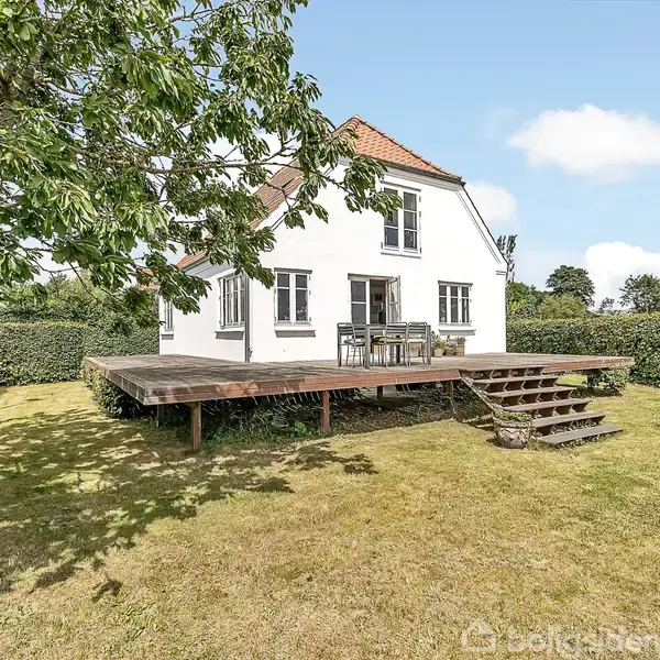 Hus med hvid facade og rødt tag står i sollys. Træterrasse foran huset med havemøbler. Græsplæne omgivet af buske og træer giver en lukket haveoplevelse.