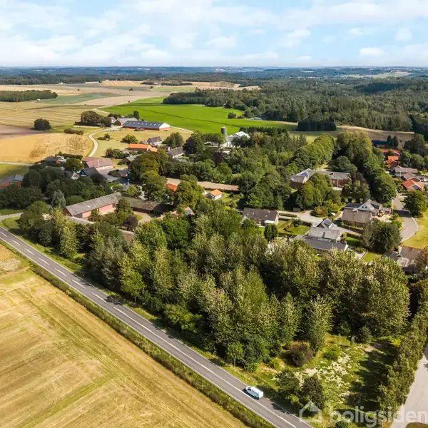 Landsby omgivet af marker og skovstrækninger, med en vej der løber gennem området. Bebyggelse består af huse og træer, landskabet strækker sig i baggrunden.