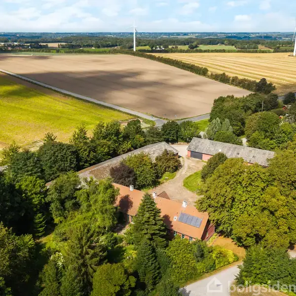 Luftfoto af gård omgivet af træer, placeret ved en landevej i landligt område med marker og vindmøller i baggrunden.