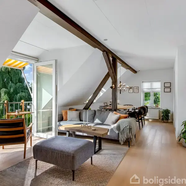 Stue med sofa og skammel, åbner ud til en balkon med udsigt til træer. Indendørs er der et spisebord, skrå vægge, træbjælker, planter og store vinduer, der slipper lys ind.