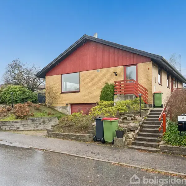 Et enfamiliehus med rødt træværk og gule mursten, placeret i en forhøjet have med trapper og buske, stående i et boligområde med en regnvåd vej foran.