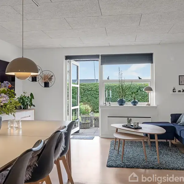 En lys stue med åbne døre til en terrasse. Et spisebord med blomster og lamper over, en blå sofa og et rundt sofabord på tæppe. Grønt hæk i baggrunden.