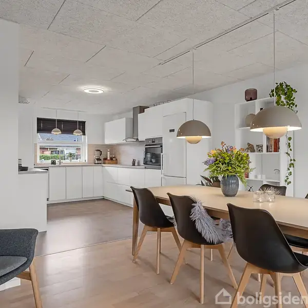 Et spisebord med stole står i en moderne, lyst køkken-alrum. Der er hængelamper over bordet, en vase med blomster og en sofa i baggrunden. Væggene er hvide og der er indbyggede hylder.